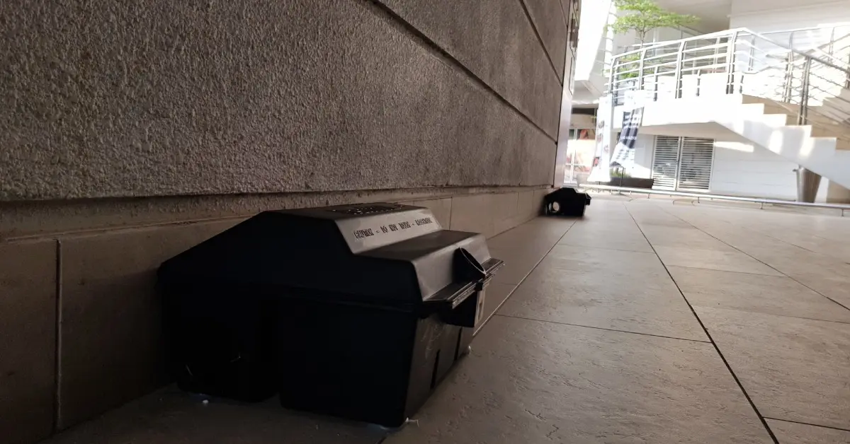 A rat trap placed in the corner of a commercial office space, under a shaded area. Another trap rests in the background.