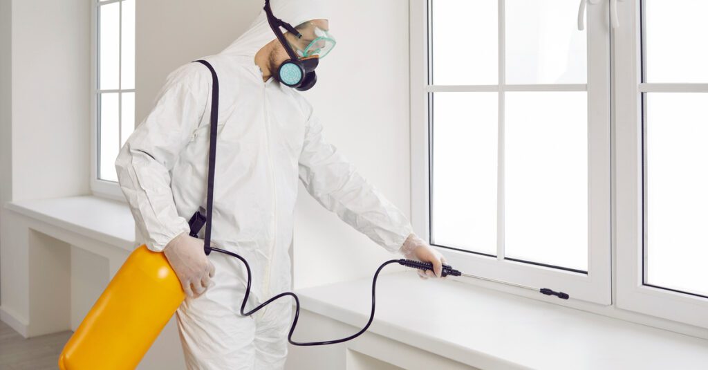 A professional exterminator, dressed in a white protective suit and respirator face covering, spraying a home with repellent.