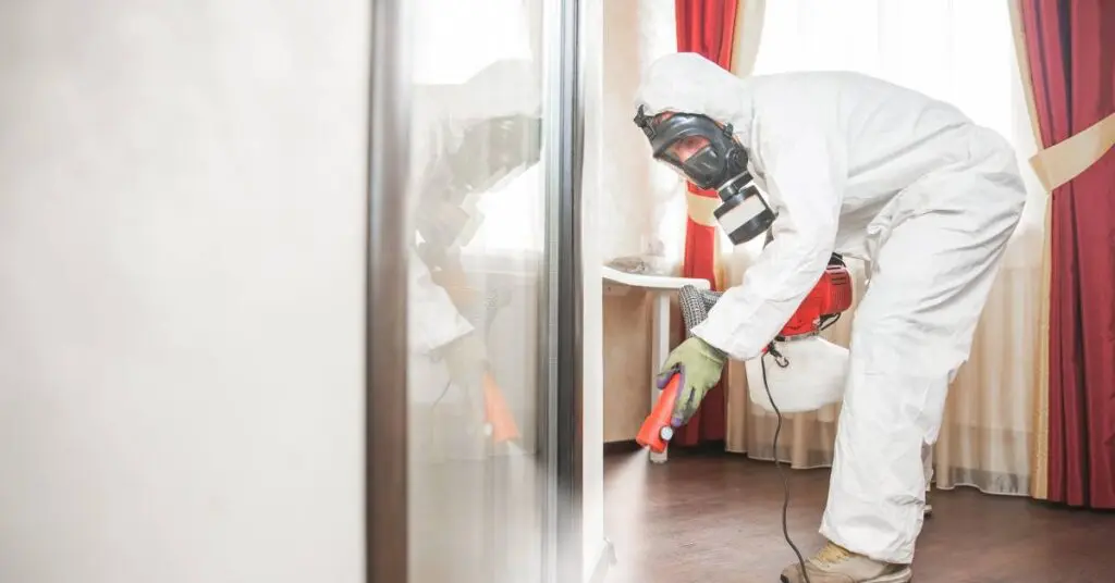 A professional rodent control specialist in white PPE sanitizes the skirting of a client's home following a pest infestation.