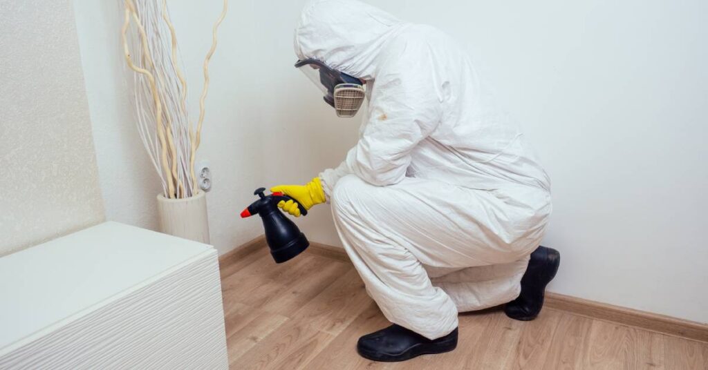 A professional pest exterminator in full protective gear spraying rodent-repellent chemicals around a client’s home.