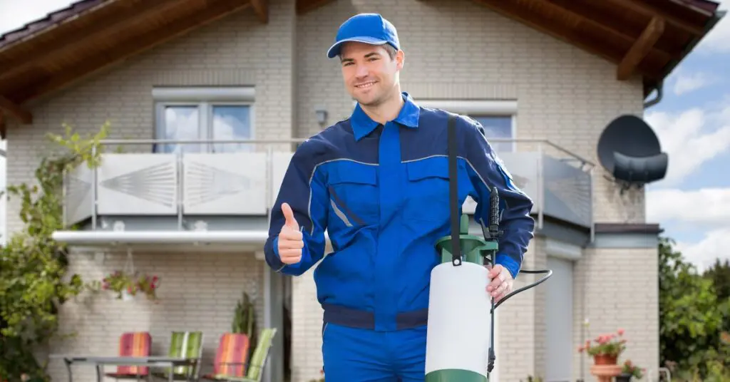 An exterminator smiles and gives a thumbs up as he stands in front of a house. He wears a blue jumpsuit.