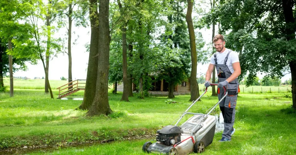 A person dressed in overalls and wearing gloves pushes a lawnmower on his lawn. A cabin sits in the background.