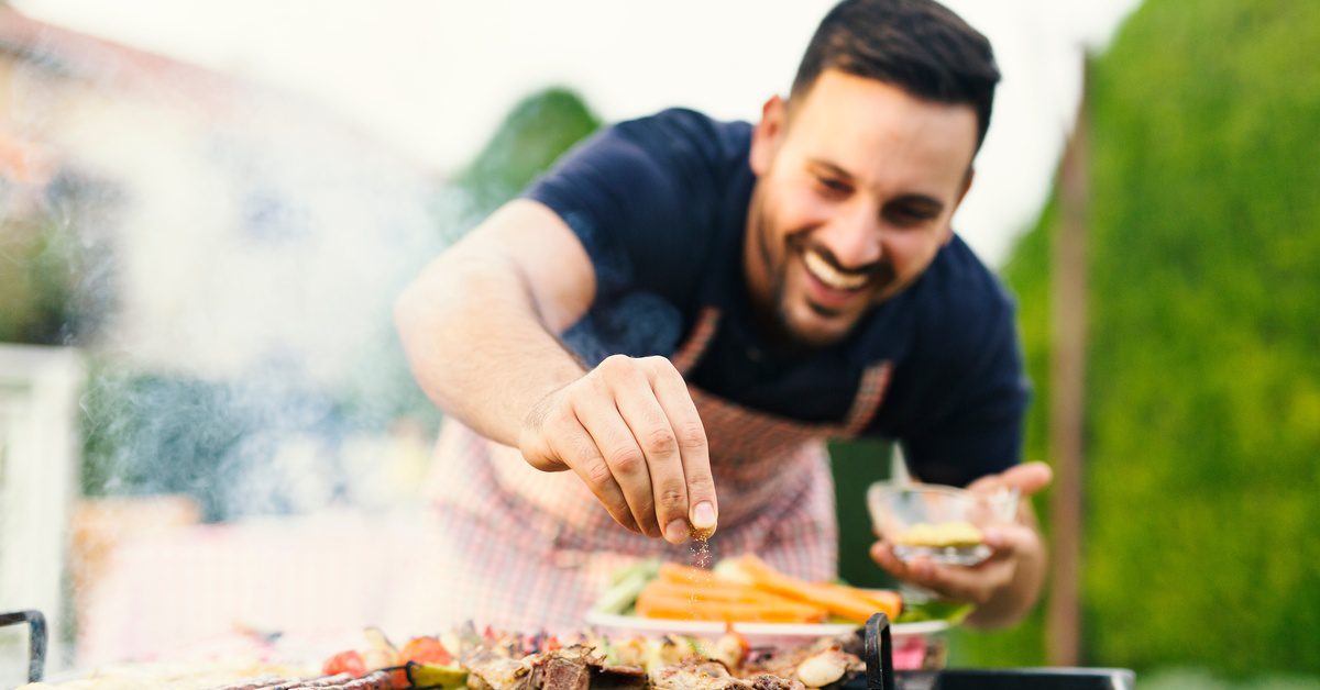 A man smiles as he sprinkles spices on meat that sits on a barbecue grill in a yard. A plate of carrots sits nearby.
