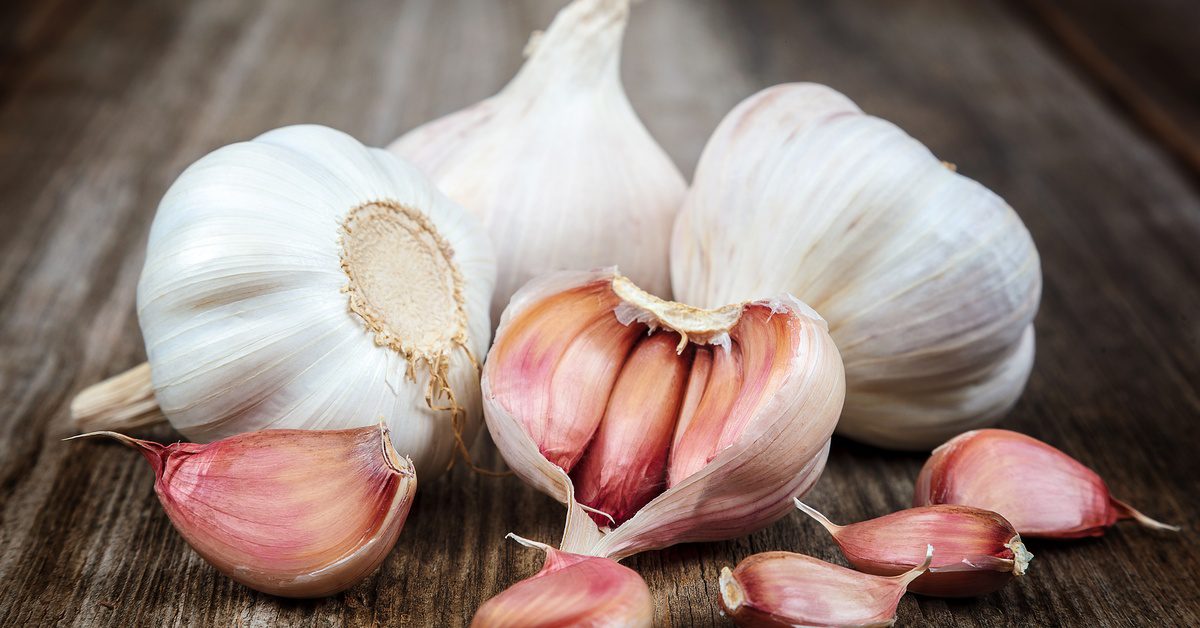 Several white garlic bulbs sit on a wooden surface. One of them is open with five cloves lying in front.