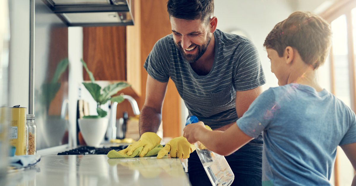 A man and little boy clean a kitchen counter together with a spray bottle and a washcloth. Both are happy.