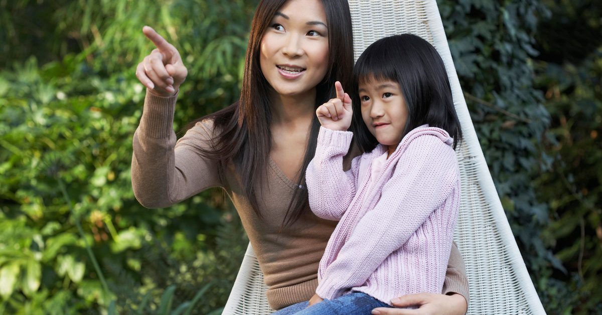 A woman and child sit on a hammock in a backyard. Both of them are pointing at something they're looking at.