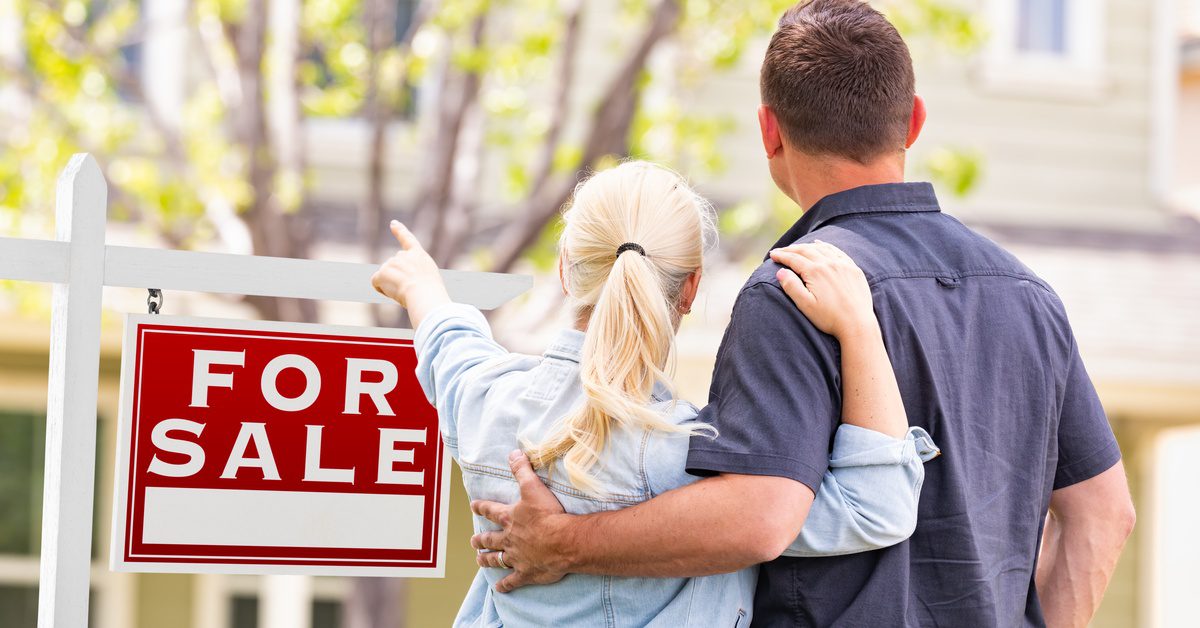 A man and a woman stand in front of a house with a tree and a "For Sale" sign. The woman points at the house.