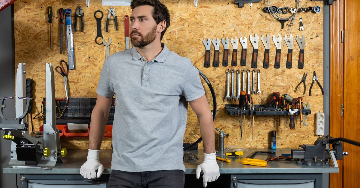 A man stands in a very organized garage while wearing white gloves. Tools hang from a board on the wall behind him.