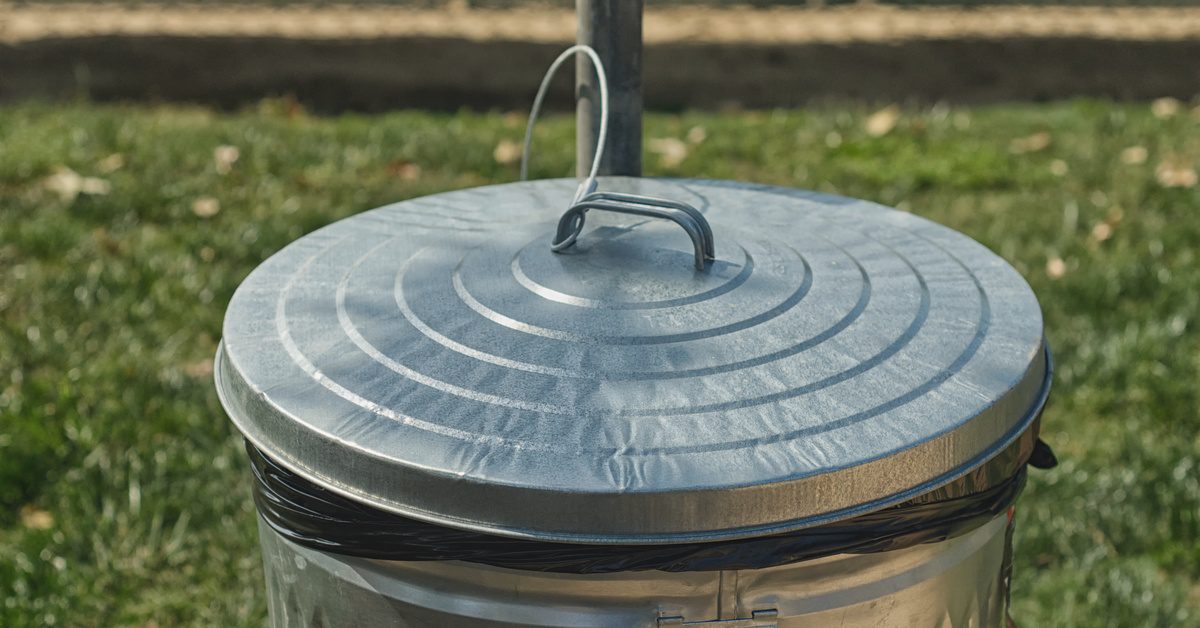 A metal trash can sits outside, with a black garbage bag and a lid. Grass and a fence are in the background.