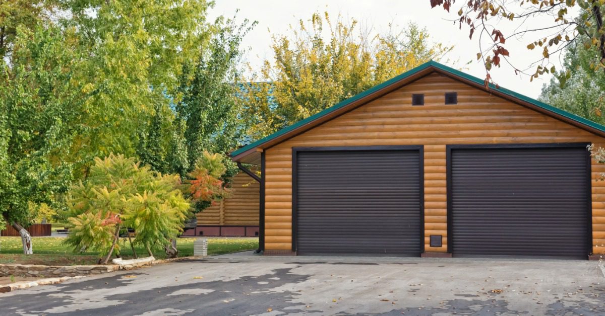 A log cabin in the woods has a garage with two doors. It stands near some trees with lots of green leaves.