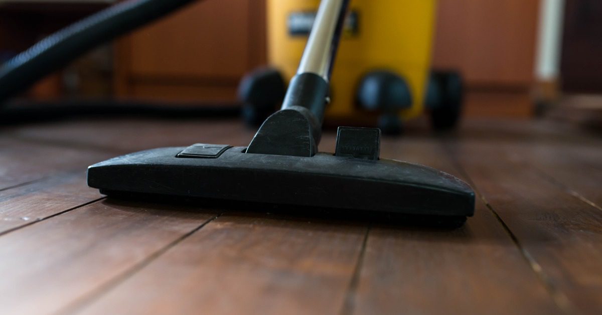 Close-up of an industrial vacuum cleaner removing dirt from a wooden floor. A yellow canister sits behind it.