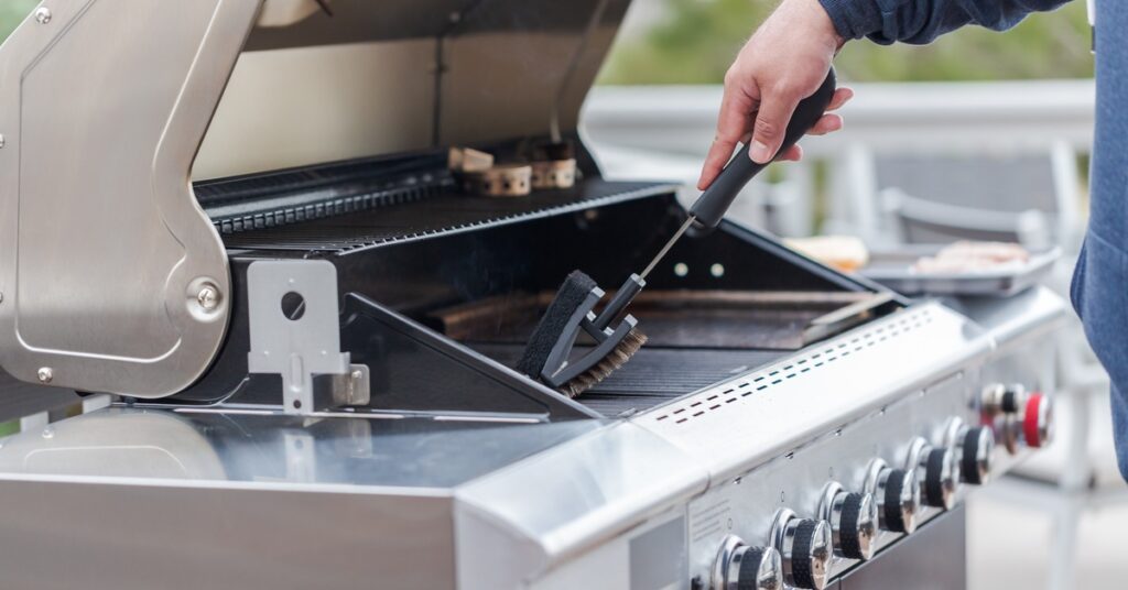 A person reaches over with a brush to scrub their backyard barbecue grill. The grill has a structure made of shiny metal.