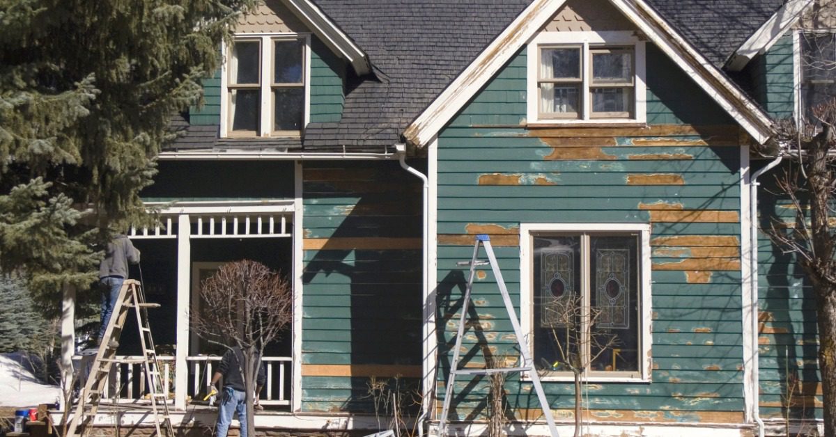 A green house's exterior features peeling paint and ladders standing outside it. A person works near the porch.