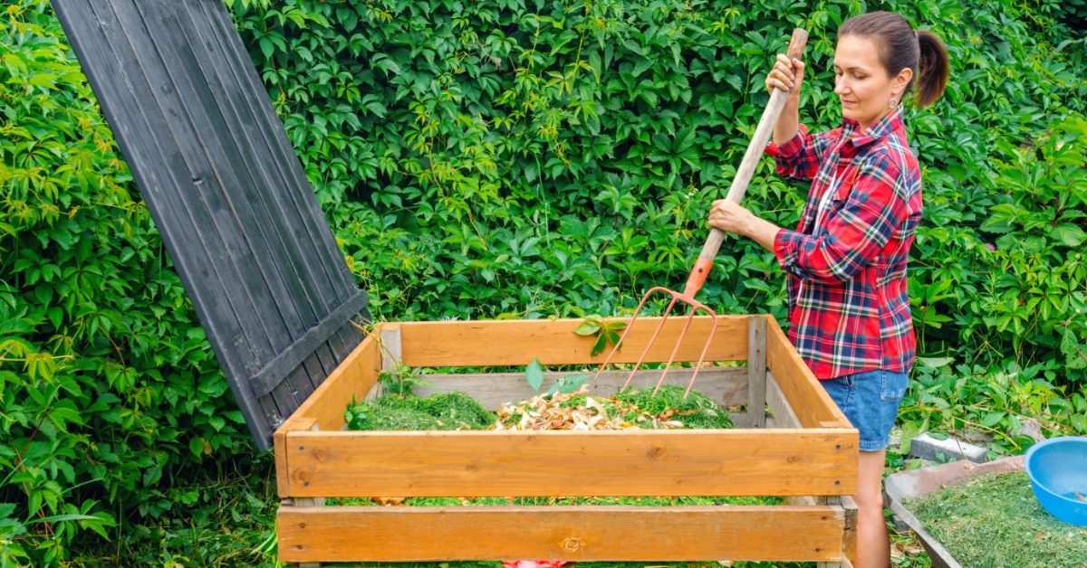 A woman in a plaid shirt stirs plant scraps in a wooden compost bin with a pitchfork. The bin has wooden sides.