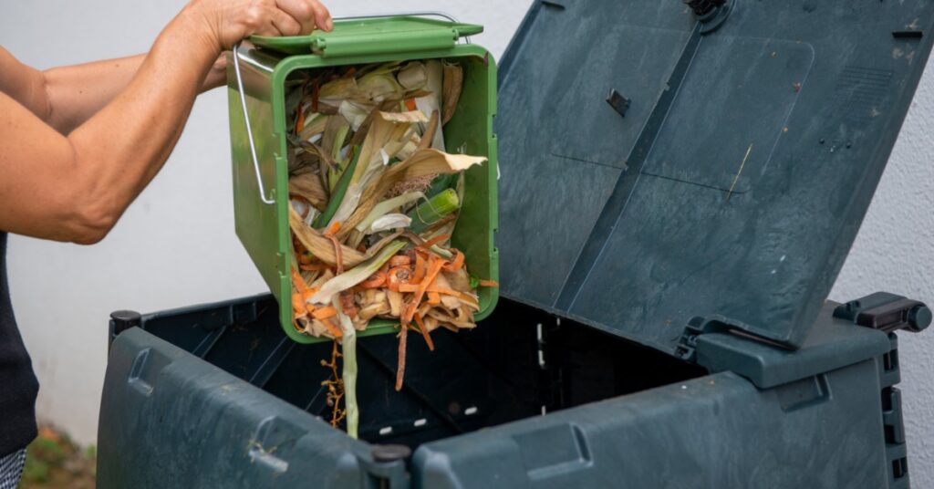 A person empties vegetable scraps from a green kitchen container into an open dark compost bin outdoors.
