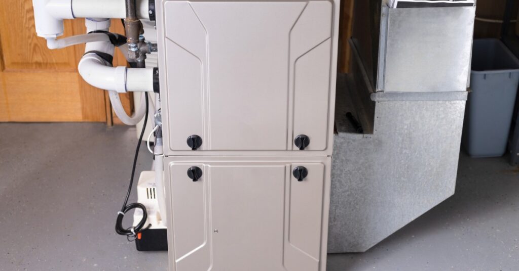 A beige gas furnace stands beside metal ductwork, white pipes and cables. It sits on a gray floor in a basement.