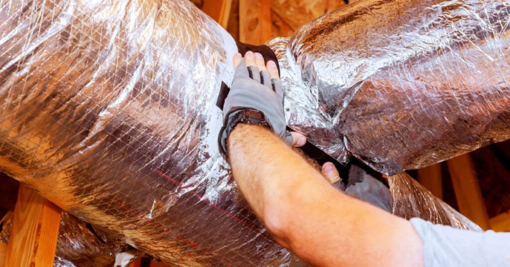 A person's gloved hands adjust reflective duct insulation between exposed wooden beams inside a wood-framed attic.