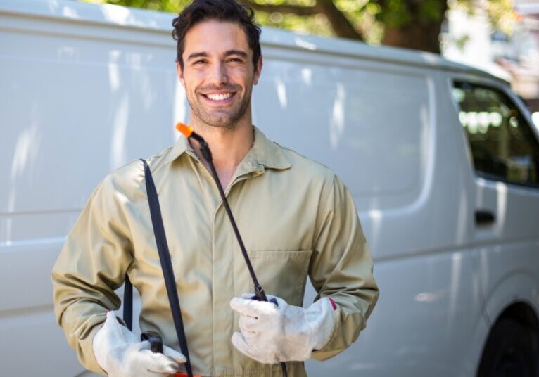 A smiling man standing outside in front of a white van. He is wearing a beige jumpsuit, gloves, and sprayer equipment.