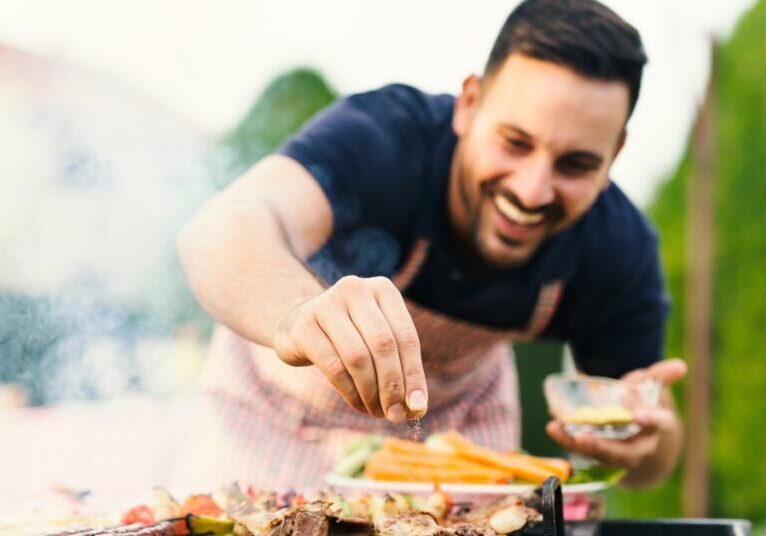 A man smiles as he sprinkles spices on meat that sits on a barbecue grill in a yard. A plate of carrots sits nearby.