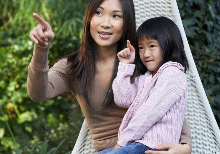 A woman and child sit on a hammock in a backyard. Both of them are pointing at something they're looking at.
