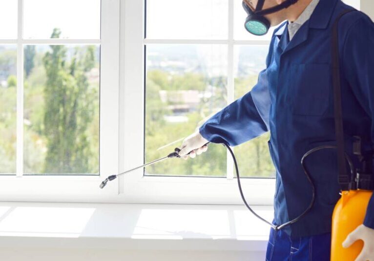 A professional exterminator in full protective gear spraying rodent repellent onto the windowsill of a client’s home.