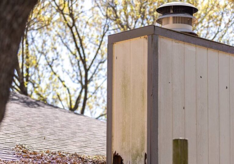 A wooden chimney stack on the roof of a home has a small opening near the base. There are leaves on the roof.