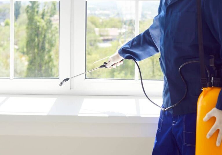 A professional rodent control specialist in a blue jumpsuit sanitizing and spraying a windowsill with pest deterrents.