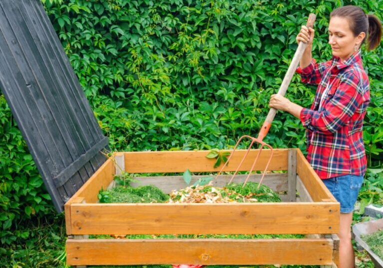 A woman in a plaid shirt stirs plant scraps in a wooden compost bin with a pitchfork. The bin has wooden sides.