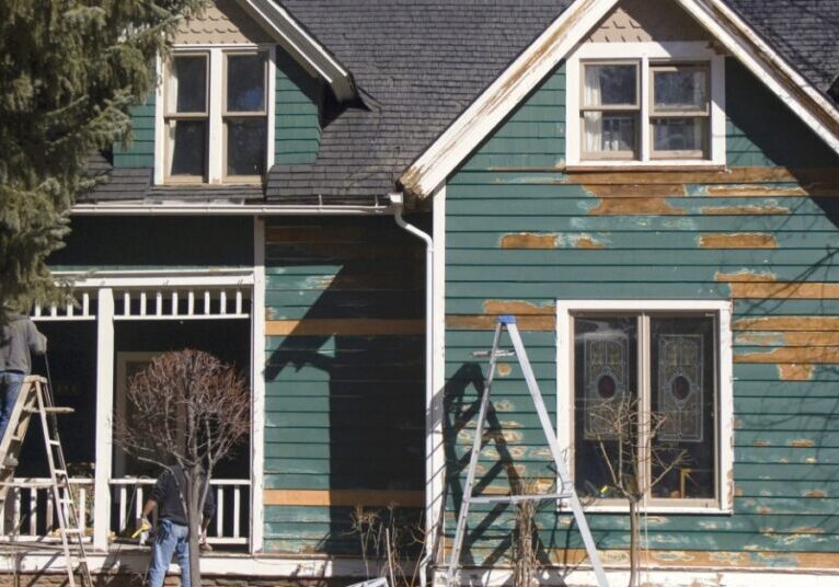 A green house's exterior features peeling paint and ladders standing outside it. A person works near the porch.